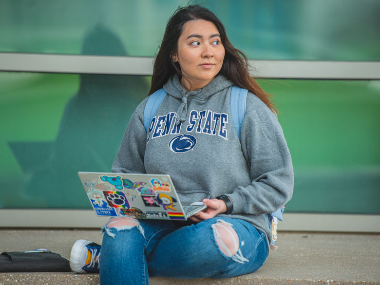 Student wearing Penn State gear works on laptop outside Gaige building