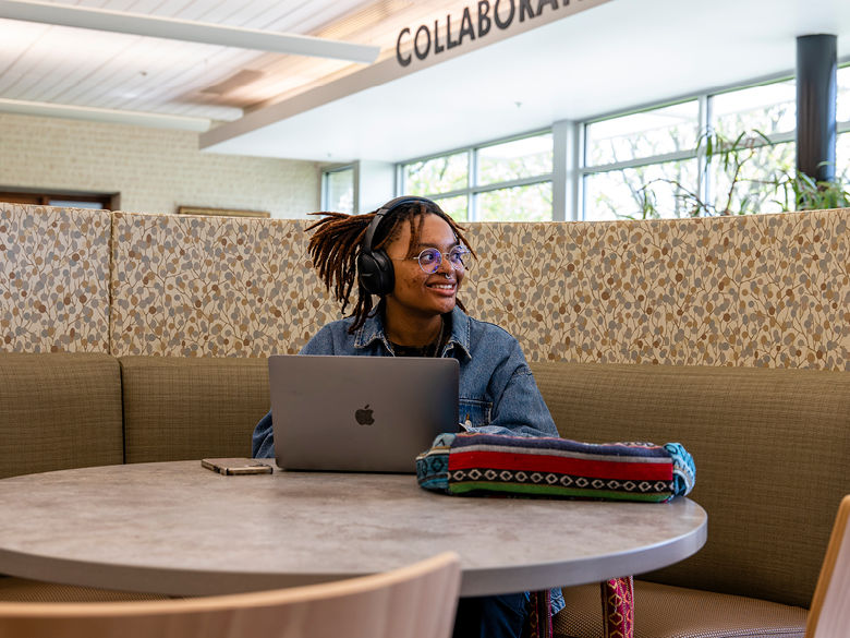Smiling female student sits with Mac laptop at table in library.